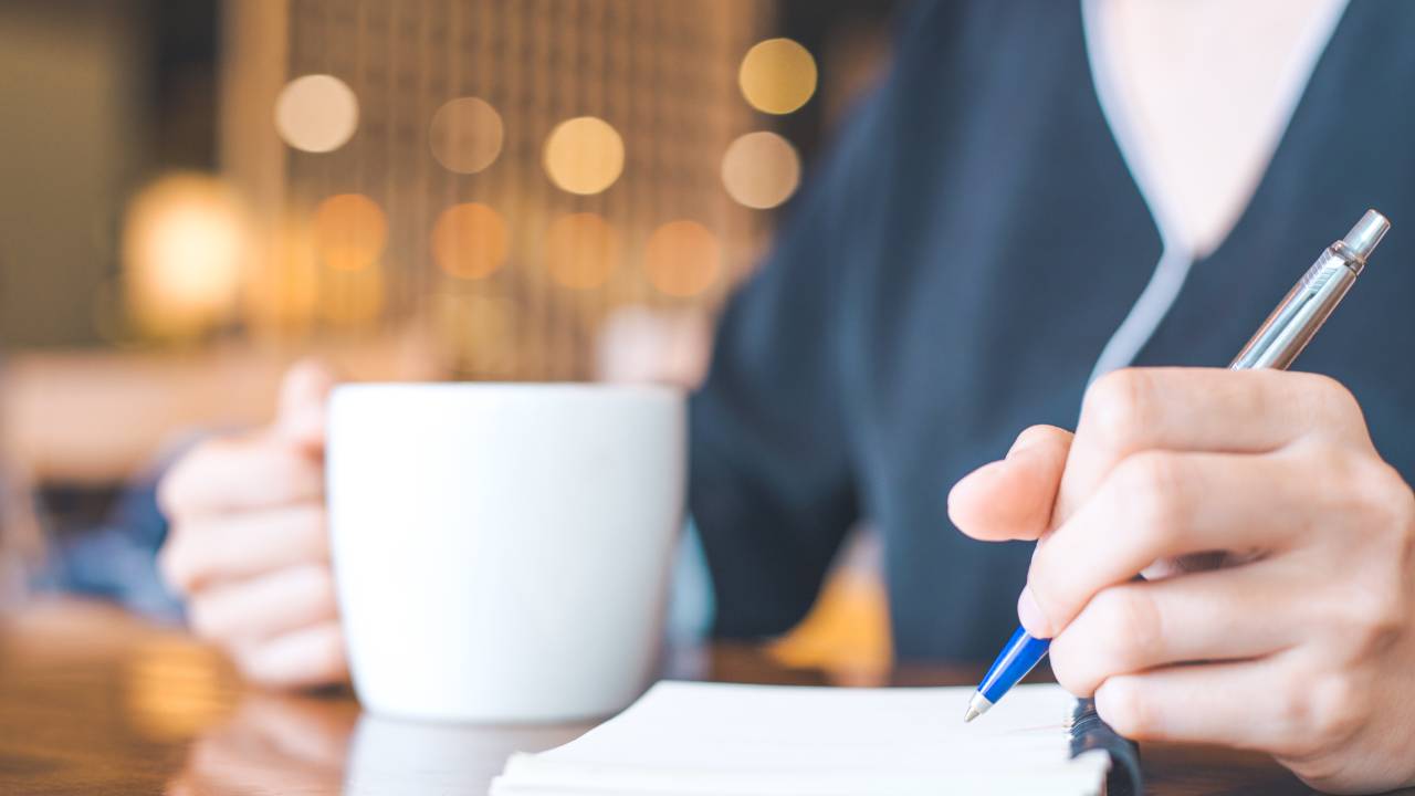 Mulher anota informações em um caderno com uma caneta em uma mão e uma xícara branca de café na outra
