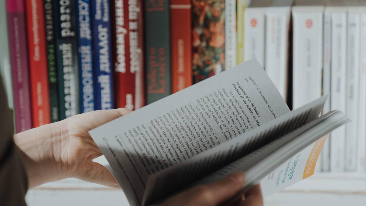Mão segurando livro em frente a uma estante.