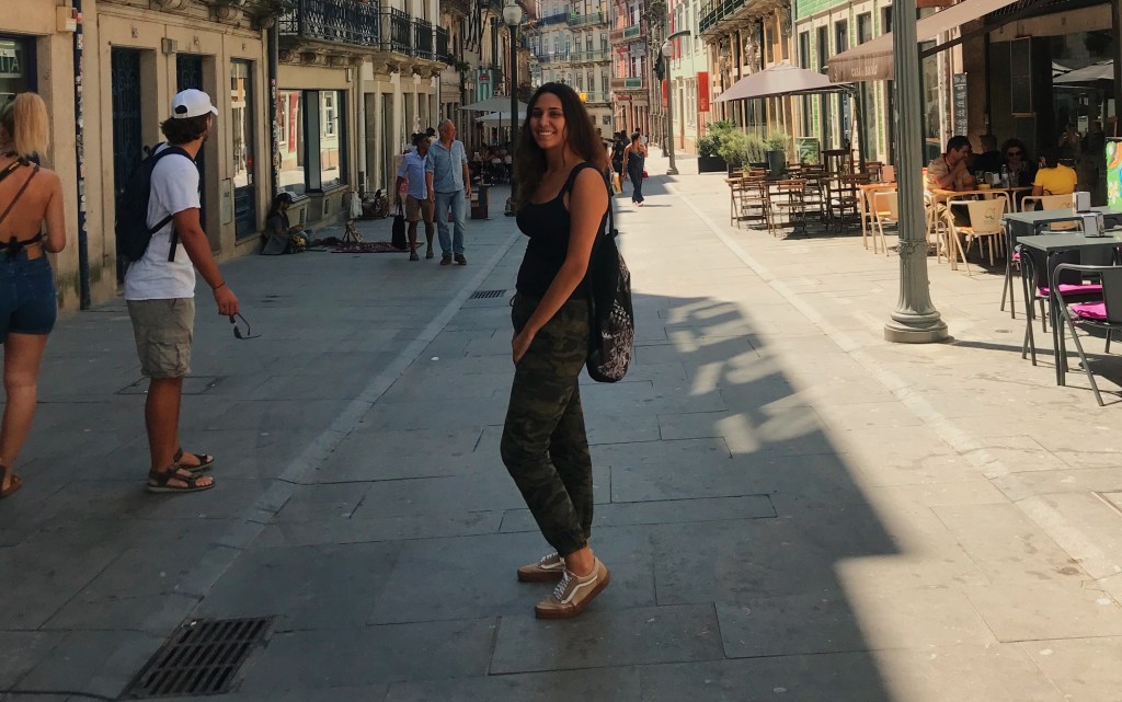 Mulher posa em uma calçada no centro de Portugal. Ela usa roupas escuras, uma mochila e apresenta um semblante feliz. Na rua, outras pessoas passam.