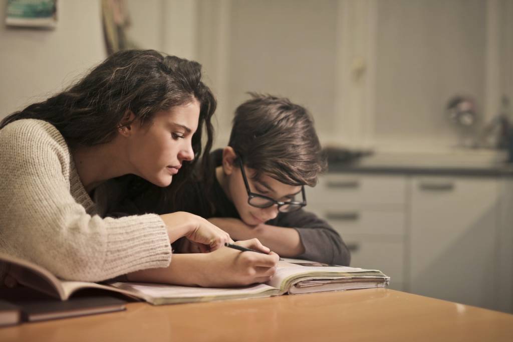 Estudo Domiciliar Mulher se apoia ao lado de um menino, que está lendo um livro. Os dois estão sentados em uma mesa dentro de um ambiente fechado.