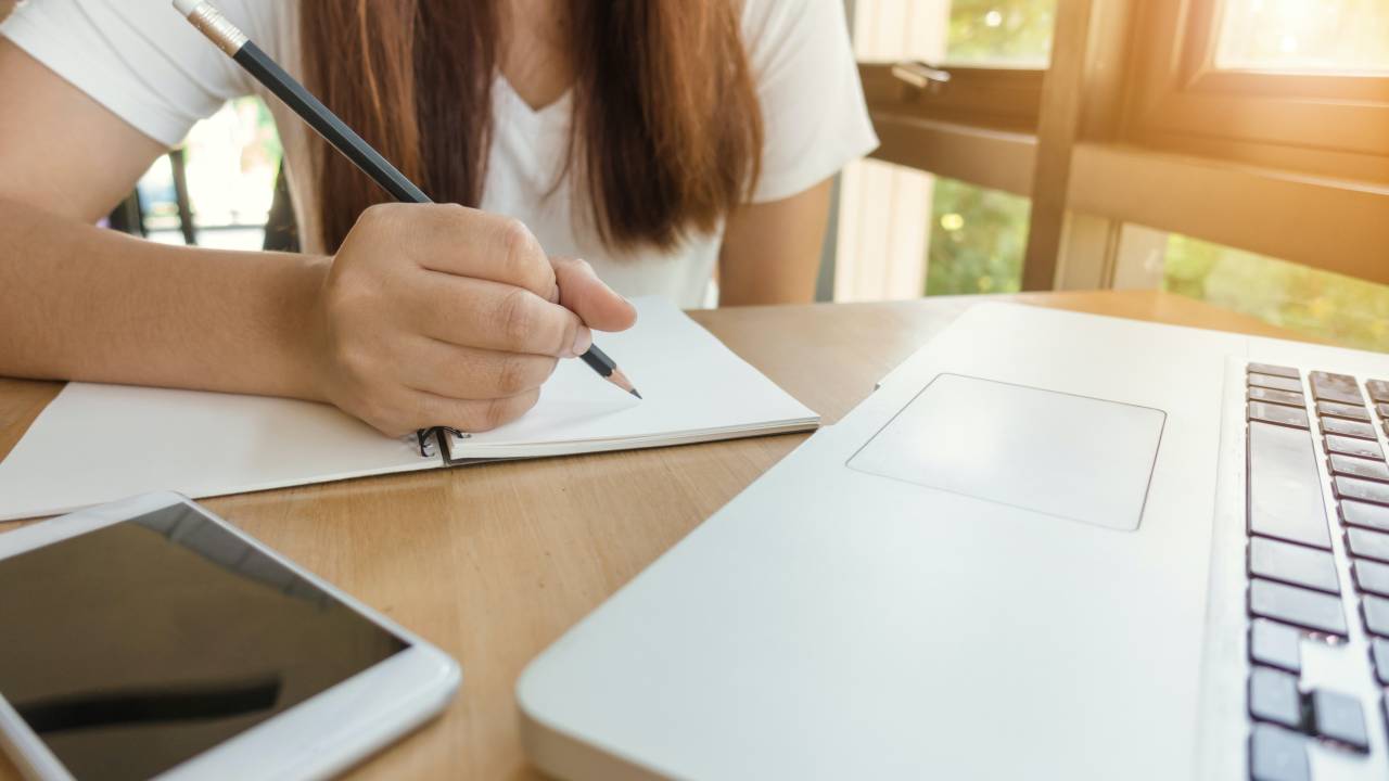 computador, celular e uma garota fazendo anotações em um papel