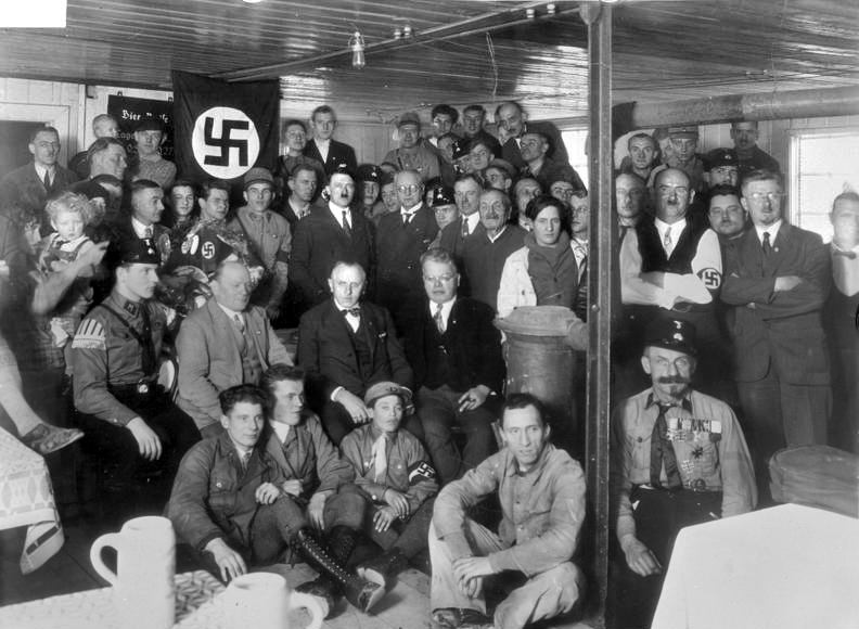 partido-nazista Grupo de homens em ambiente interno, há uma bandeira nazista ao fundo