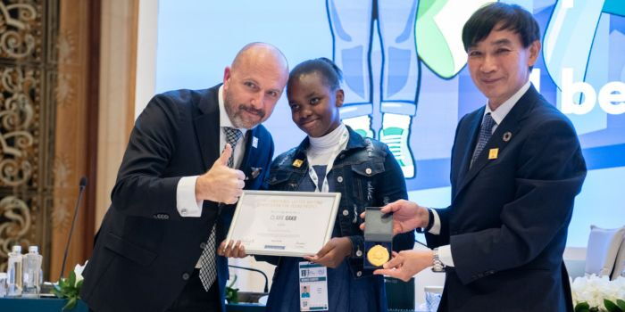claire_01 Jovem de 13 anos posa com seu certificado ao lado de dois representantes da União Postal Universal em palco de cerimônia na Suíça