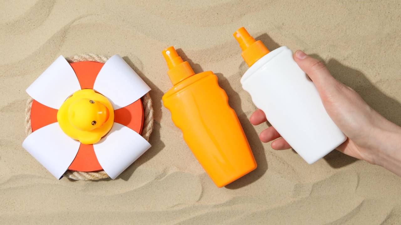 Na areia, uma mão pega uma embalagem de protetor solar branca, ao lado de outra laranja. Há ainda um pato amarelo de borracha
