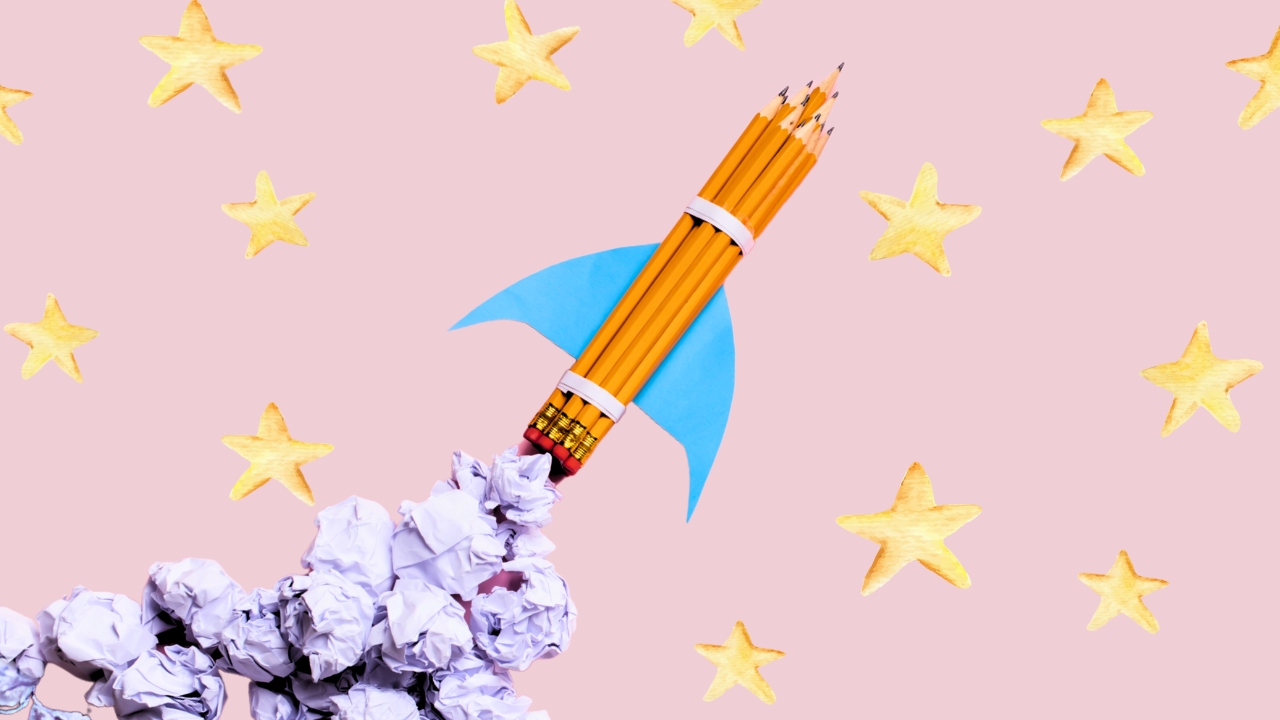 Conjunto de lápis de escrever unidos por 2 elásticos, com duas asas de cartolina coladas na lateral, simulando a silhueta de um foguete em lançamento. No fundo, um papel rosa claro, com estrelas amarelas desenhas