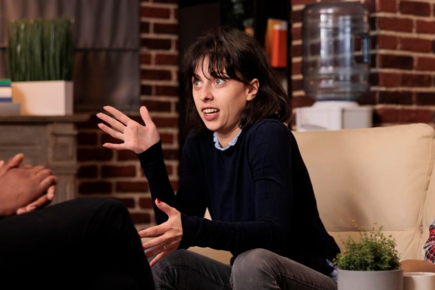 balbuciar Mulher falando, gesticulando, confusa. Está sentada em sofá conversando com outros dosi homens.