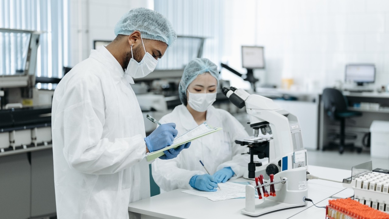 Dois cientistas, um homem e uma mulher, em um laboratório. O homem, à esquerda, anota em uma prancheta. A mulher, à direita, escreve em uma folha, com um microscópio ao lado. Ambos usam jalecos brancos, toucas, máscaras e luvas azuis, em um ambiente claro com equipamentos de pesquisa.